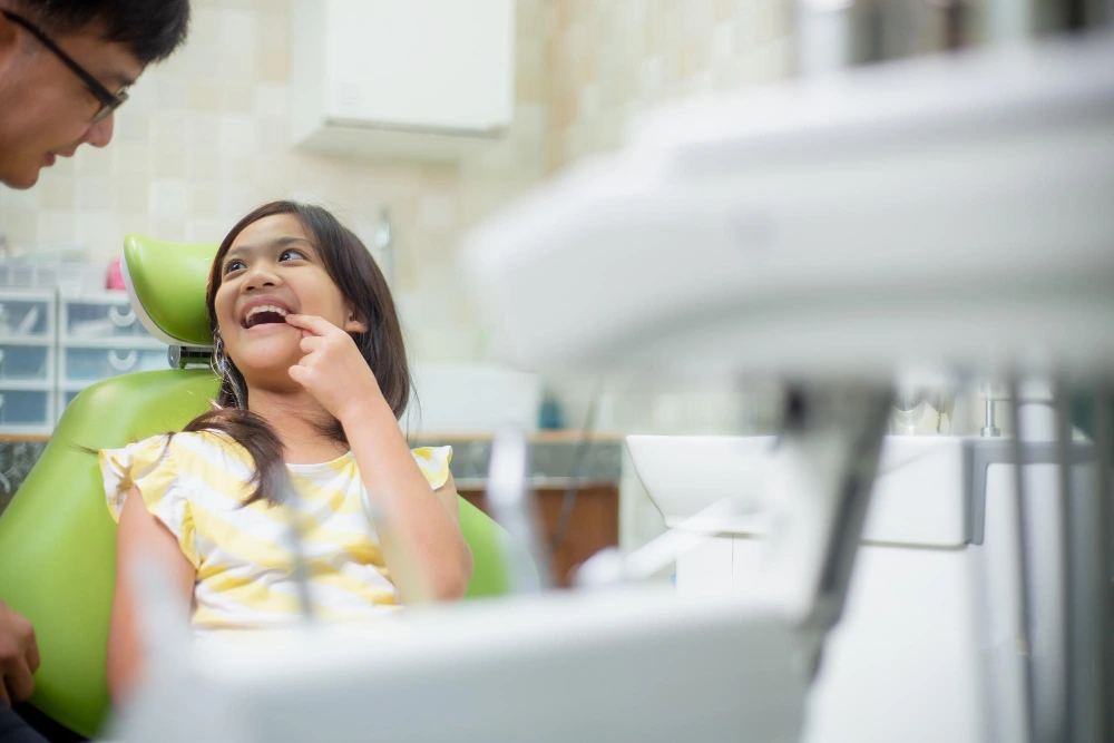 Young girl in a green dental chair smiling as a dentist talks with her in a clinic visit aftermath?