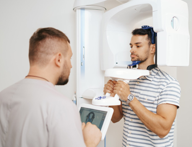 Man undergoing a panoramic dental X-ray; technician adjusts the machine while holding a control tablet nearby.