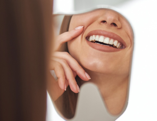 Smiling woman glancing into a small handheld mirror, teeth visible in the reflection