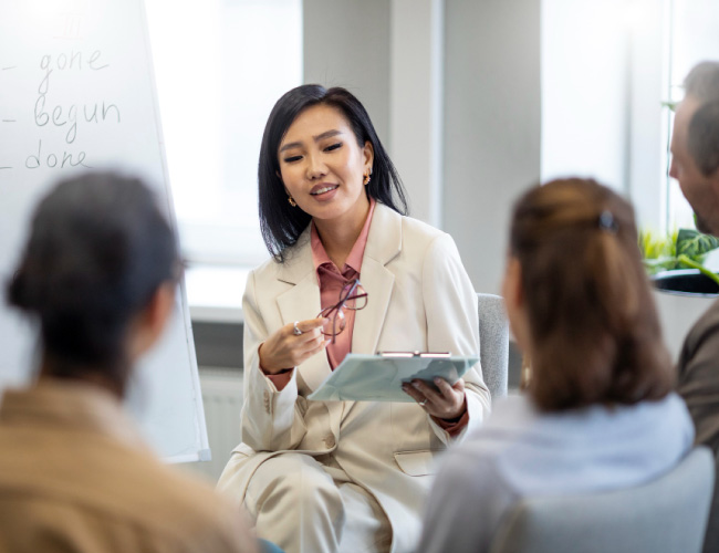 Businesswoman in a light suit leads a small meeting, holding a clipboard and glasses as colleagues listen.