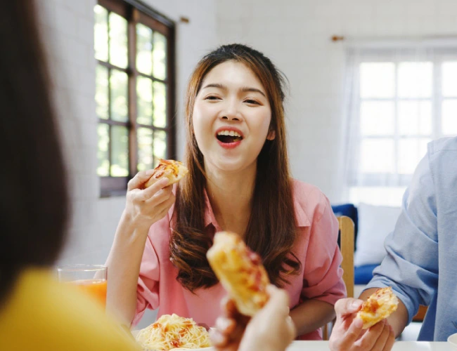 Young woman in a pink shirt smiles while taking a bite of pizza at a dining table with friends indoors.