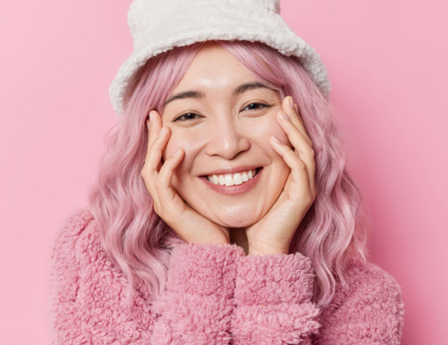 Smiling woman with pastel pink hair, wearing a white fuzzy hat and pink fleece sweater against a pink backdrop.
